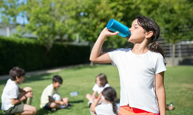 Child drinking from a refillable water bottle outdoors, highlighting hydration and eco-friendly habits for students.