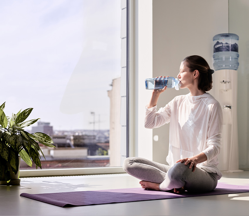 Frau trinkt entspannt aus Wasserflasche auf Yogamatte vor einem Wasserspender