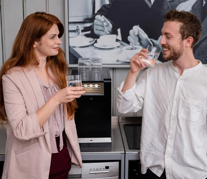 Zwei Personen stehen vor einem Leitungsgebundenen Wasserspender und trinken Wasser während sie sich unterhalten