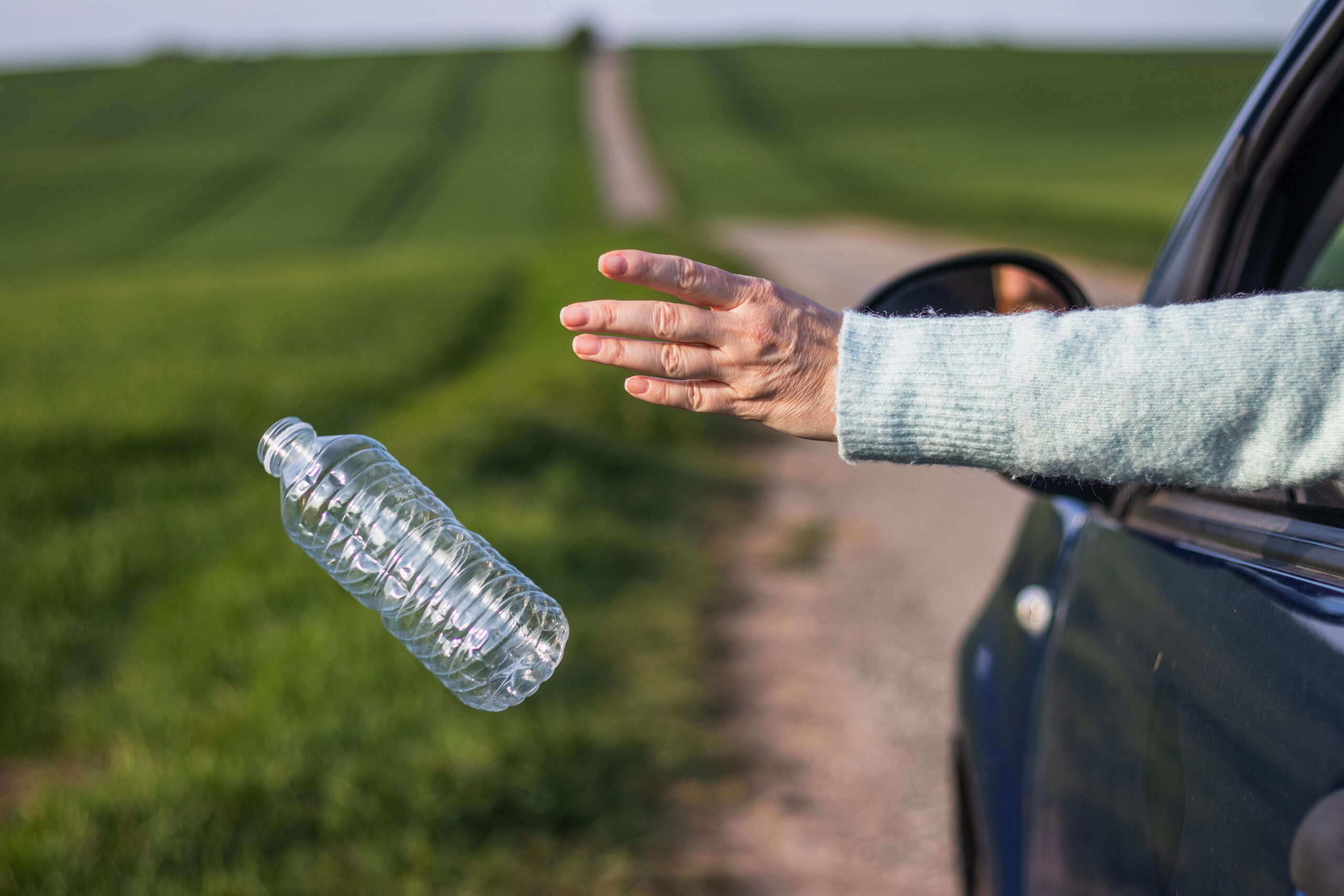 Person wirft Plastikflasche aus dem Auto in grüne Landschaft