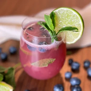 Erfrischender Sommerdrink mit Heidelbeeren, Limettenscheibe, frischer Minze und Eiswürfeln in einem Glas auf einem Holztisch, im Hintergrund verstreute Heidelbeeren und ein weißes Tuch.