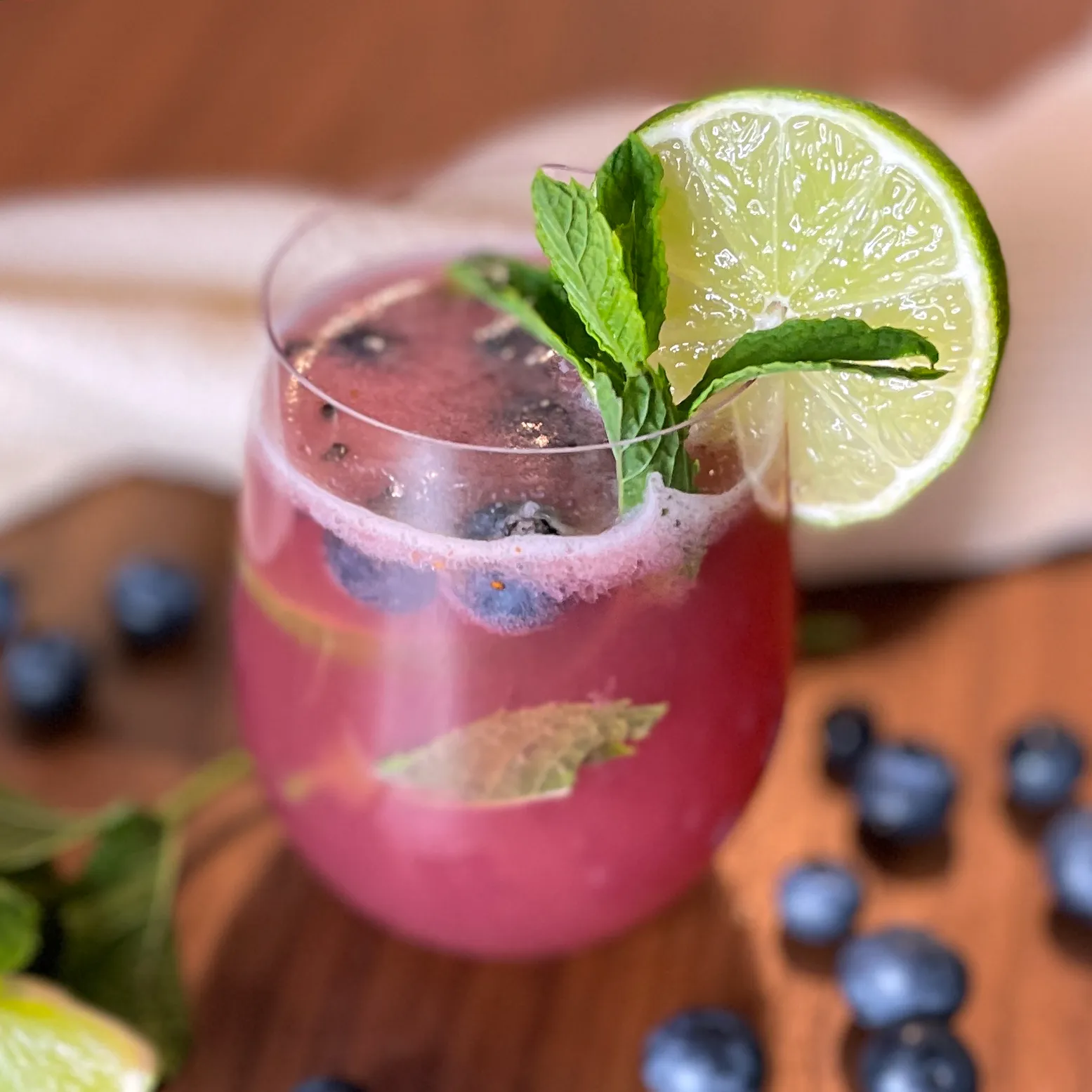 Erfrischender Sommerdrink mit Heidelbeeren, Limettenscheibe, frischer Minze und Eiswürfeln in einem Glas auf einem Holztisch, im Hintergrund verstreute Heidelbeeren und ein weißes Tuch.