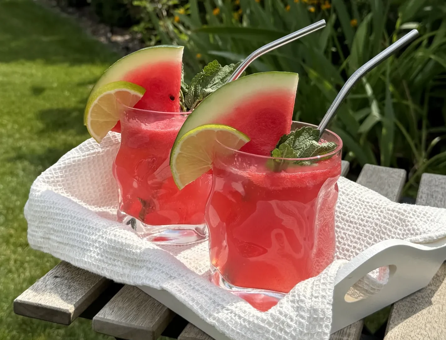 Zwei Gläser mit leuchtend rotem Sommergetränk aus Wassermelone, garniert mit Limettenscheiben, frischer Minze und Edelstahlstrohhalmen, serviert auf einem weißen Tablett im Garten mit grüner Pflanzenkulisse.