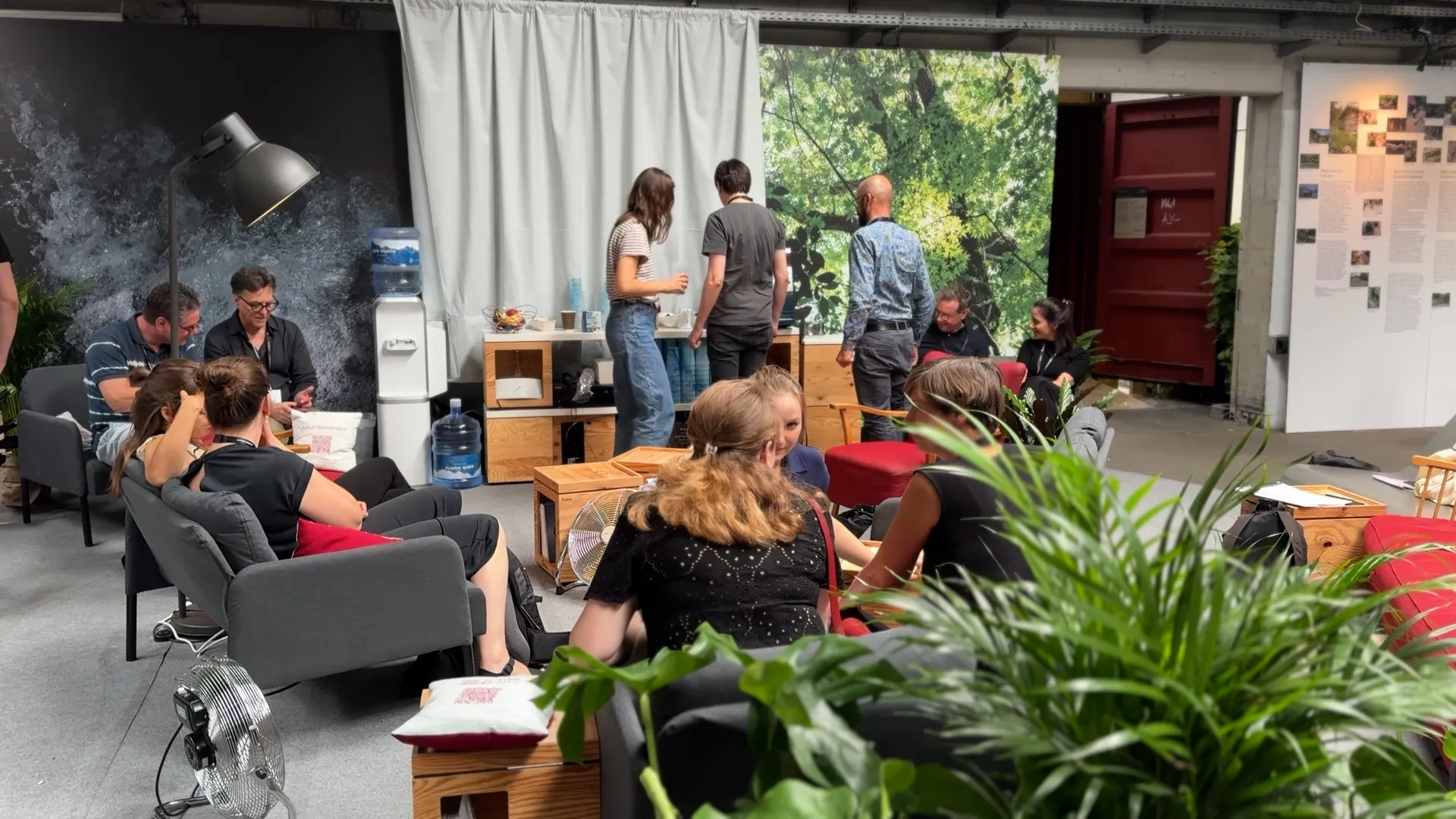 Moderne Lounge mit Menschen in Gesprächsrunden und entspannter Atmosphäre, im Hintergrund ein Wasserspender mit Culligan Kanister und Holzregalen mit Bechern und Obstschale.