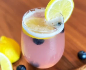 Glas mit pinker Blaubeerlimonade, frischen Heidelbeeren mit Zitronenscheiben, dekorativ serviert auf Holzuntergrund mit ganzen Zitronen und Heidelbeeren.