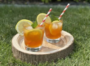 Zwei Gläser mit orangefarbenem Sommergetränk, garniert mit Zitronenscheiben, Minze und rot-weißen Papierstrohhalmen, serviert auf einem runden Holzbrett auf einer grünen Wiese mit blühenden Gartenpflanzen im Hintergrund.