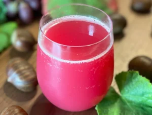 Glas mit leuchtend rotem Sturm-Spritzer, umgeben von Kastanien, Weintrauben und herbstlich gefärbten Blättern auf einem Holztisch.