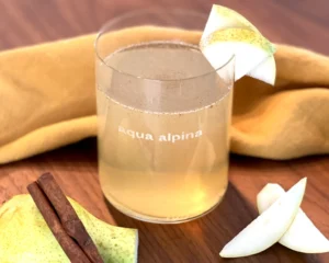 Herbstliches Getränk mit Birne und Zimt in einem aqua alpina Glas, dekoriert mit Birnenspalten, daneben frische Birne, Zimtstangen und ein gelbes Tuch auf Holzuntergrund.